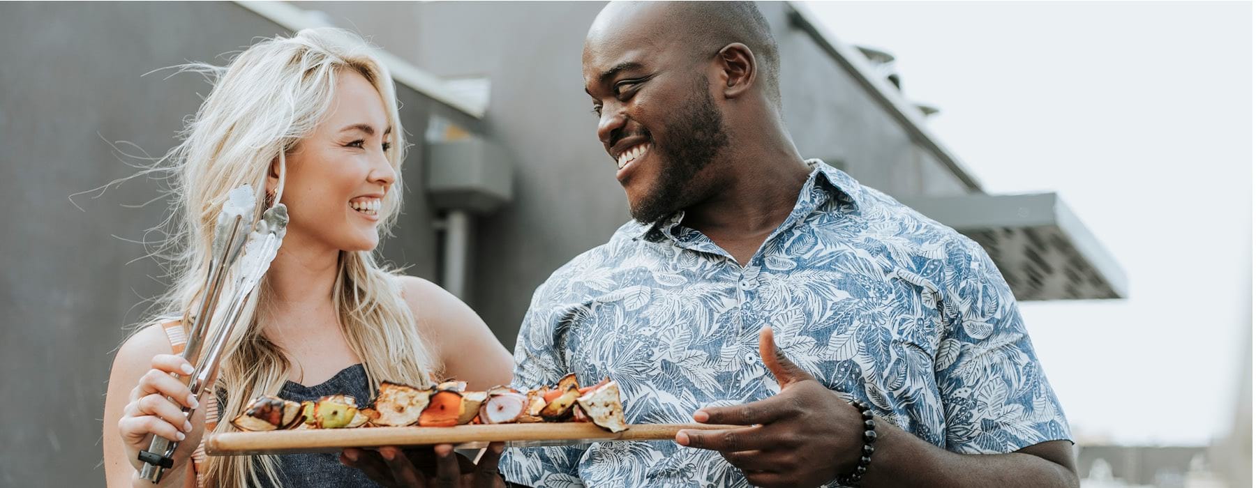 man and woman smile at each other while holding a cutting board of kabobs and grilling tongs